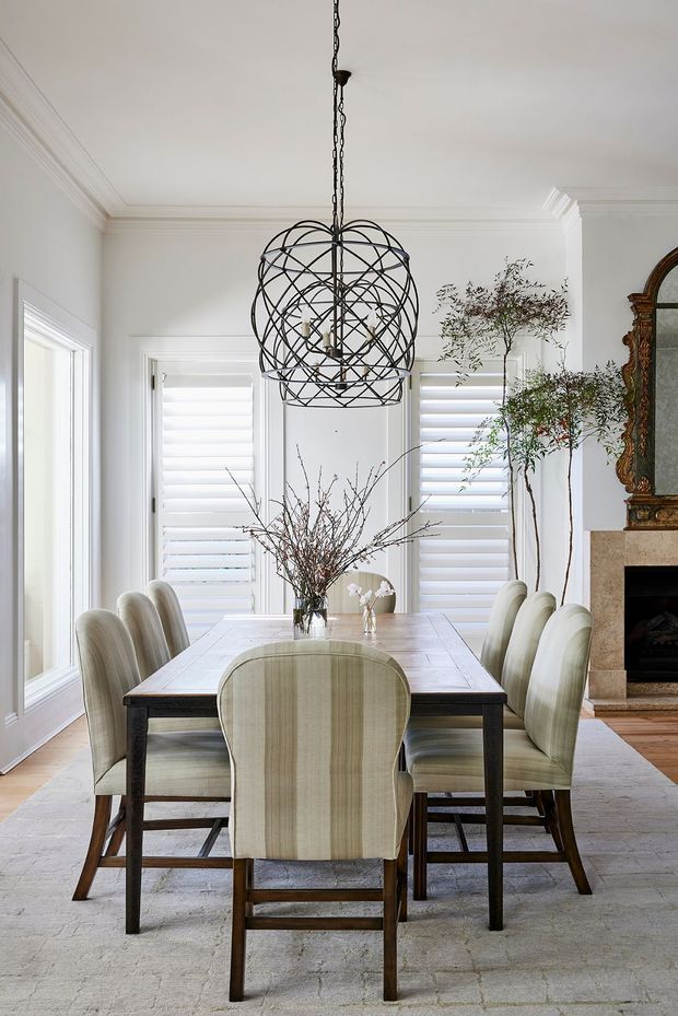 Bright, elegant dining room with a large rectangular timber dining table surrounded by upholstered dining chairs. White walls, detailed crown moulding, plantation shutters, a sculptural black metal spherical pendant light, pale flooring, and a traditional ornate fireplace surround create a refined transitional interior. Natural branches in vases add an organic decorative element.