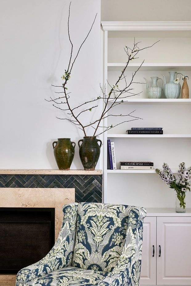 A refined living room vignette featuring a patterned upholstered armchair in the foreground, a fireplace with dark green herringbone tile surround and pale stone mantel, and built-in white shelving and cabinetry. The shelves display ceramic vessels, books, and a vase of flowers, while large bare branches in earthenware vessels add an organic decorative element.