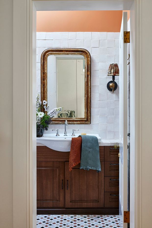 A compact bathroom viewed through an open doorway, featuring a dark timber vanity with a white basin, chrome tapware, white textured wall tiles, patterned geometric floor tiles, and a large antique-style framed mirror. An orange-painted upper wall and colourful towels add warmth and character, with a vintage wall light beside the mirror.