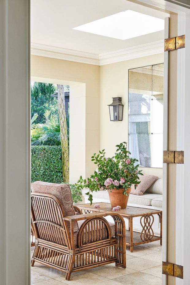 Bright, elegant enclosed entry or sunroom with cream walls, ornate white crown moulding, a large skylight, tiled floor, and a full-height mirror. Wicker and rattan armchairs surround a small wooden table with a potted flowering plant, while a cushioned built-in bench sits against the wall. An open doorway reveals lush garden greenery outside, and decorative bronze-toned door hardware and a wall lantern add traditional detail.