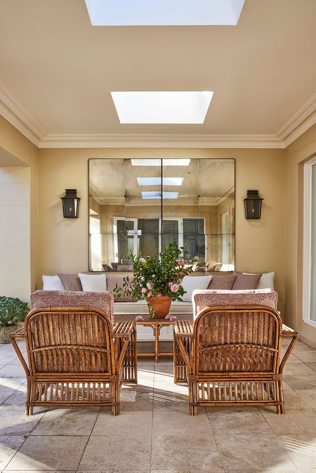 A bright, formal sitting room with warm beige walls, ornate crown moulding, and a large mirrored feature wall. Two woven rattan lounge chairs face a pale upholstered sofa, with a glass-topped coffee table, floral arrangement, wall lanterns, and tiled flooring. Skylight-like ceiling openings and the mirror create a light-filled, spacious feel.