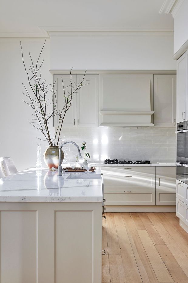 Bright, refined white kitchen with painted Shaker-style cabinetry, a large marble-look island with an undermount sink and curved chrome faucet, white subway-tile backsplash, integrated rangehood, gas cooktop, and built-in oven. Wide pale timber floorboards, ornate ceiling cornices, and delicate branch styling create a light, classic atmosphere.