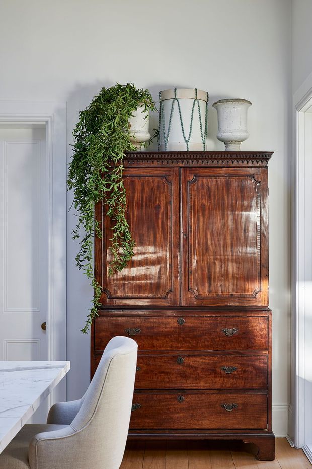 A bright, refined interior featuring a tall antique-style dark timber secretary cabinet with paneled doors and multiple drawers. The cabinet is styled with ceramic planters and a trailing green plant, beside a white upholstered chair and a pale stone-topped surface. White paneled walls, trim, and doors create a classic backdrop, with warm timber flooring and strong natural light from the right.