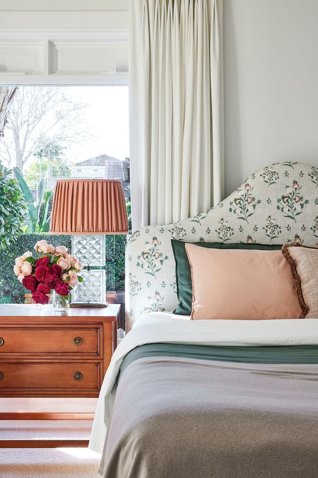 A refined, traditionally styled bedroom with a floral upholstered bedhead, layered blush, green and neutral bedding, a timber bedside chest, pleated table lamp, full-height cream curtains and a large bright window. The room combines classic detailing with soft botanical textiles and warm natural light.