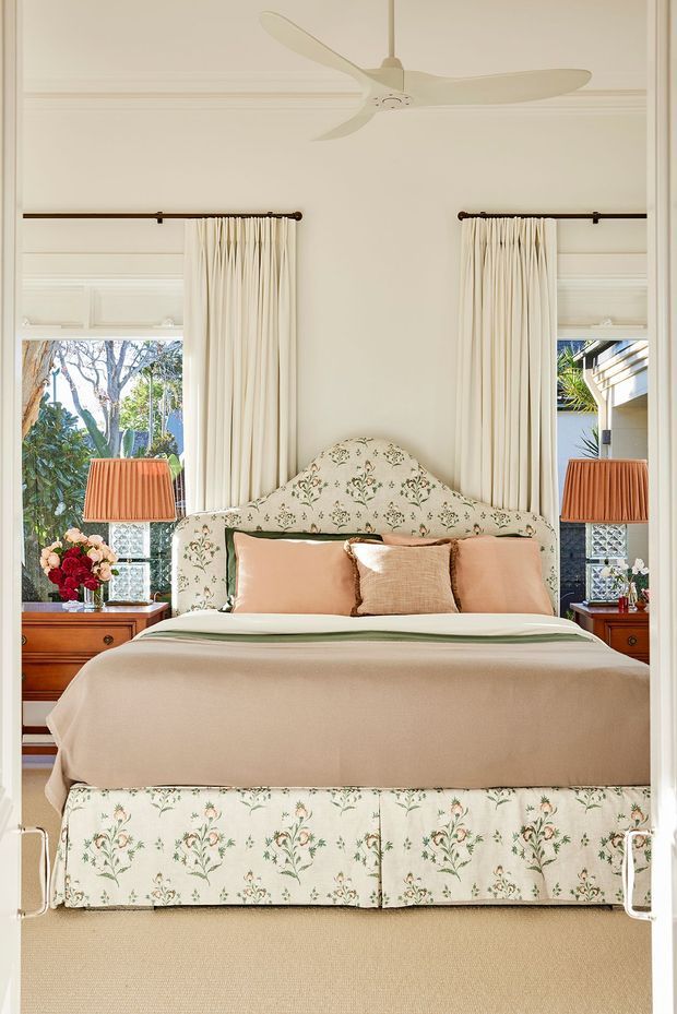 A bright, symmetrical bedroom with a floral upholstered bed and matching floral headboard and valance. The bed is dressed in cream and blush-toned linens with decorative pillows, flanked by timber bedside tables, pleated table lamps, and tall cream curtains over paired windows. White decorative wall panelling, a ceiling fan, and a neutral carpet complete the refined traditional interior.