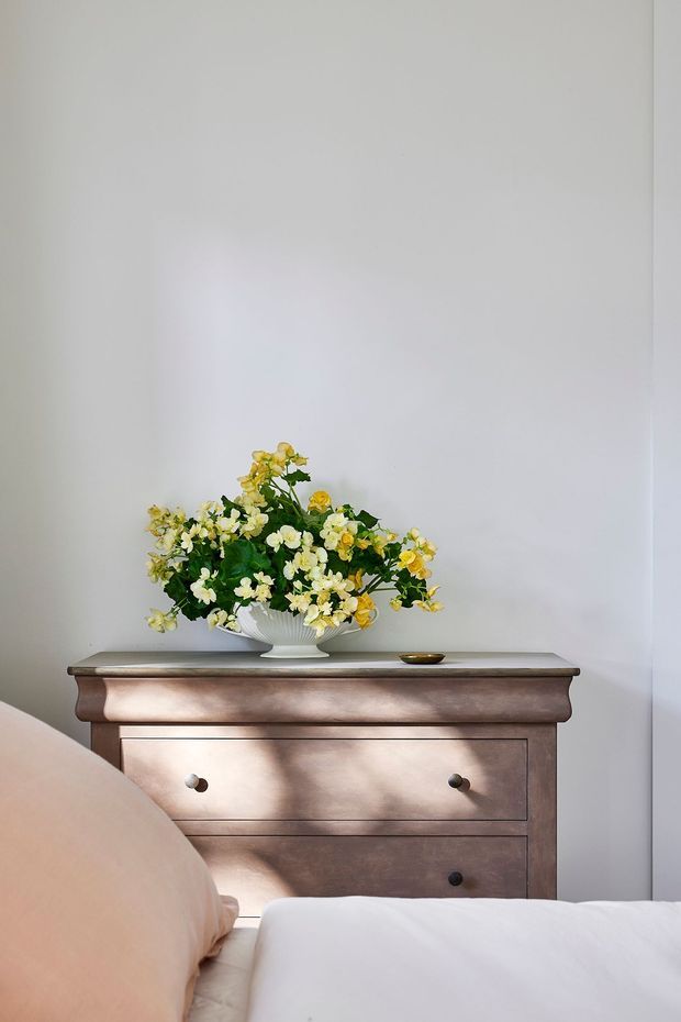 A softly lit bedroom vignette featuring a traditional dark wood two-drawer dresser against a plain light wall. A white ceramic bowl filled with yellow and white flowers sits on the dresser, with the edge of a bed, pale peach pillow, and white bedding visible in the foreground.