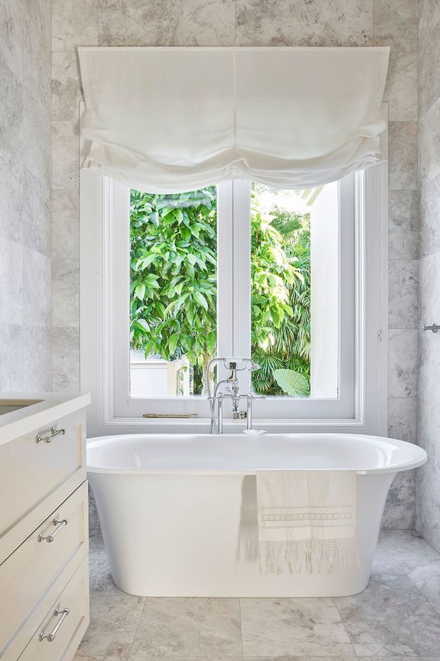 Bright, elegant bathroom featuring a freestanding white bathtub positioned beneath a large white-framed window. The walls and floor are clad in pale grey veined stone-look tiles, with a light cream built-in vanity at left, chrome tapware, and a white fabric Roman blind. Lush greenery is visible outside the window.