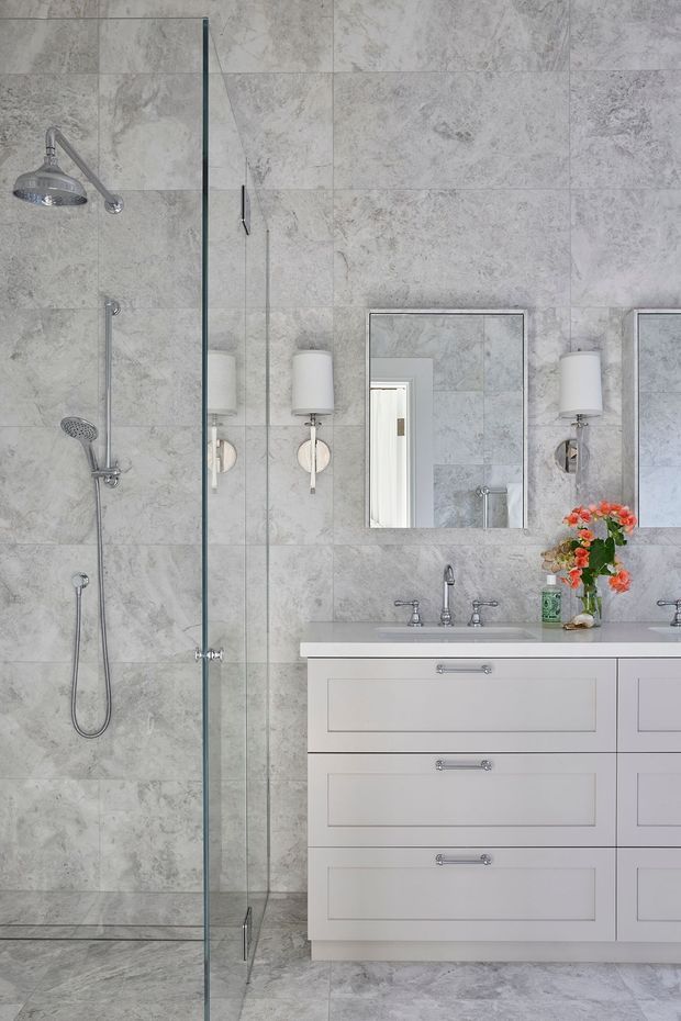 A bright, refined bathroom with a large frameless glass walk-in shower, handheld and overhead chrome shower fittings, grey marble-look wall and floor tiles, and a white double vanity with recessed-panel drawers and a pale stone benchtop. Rectangular mirrors and matching wall sconces sit above the vanity, with a small floral arrangement adding colour.