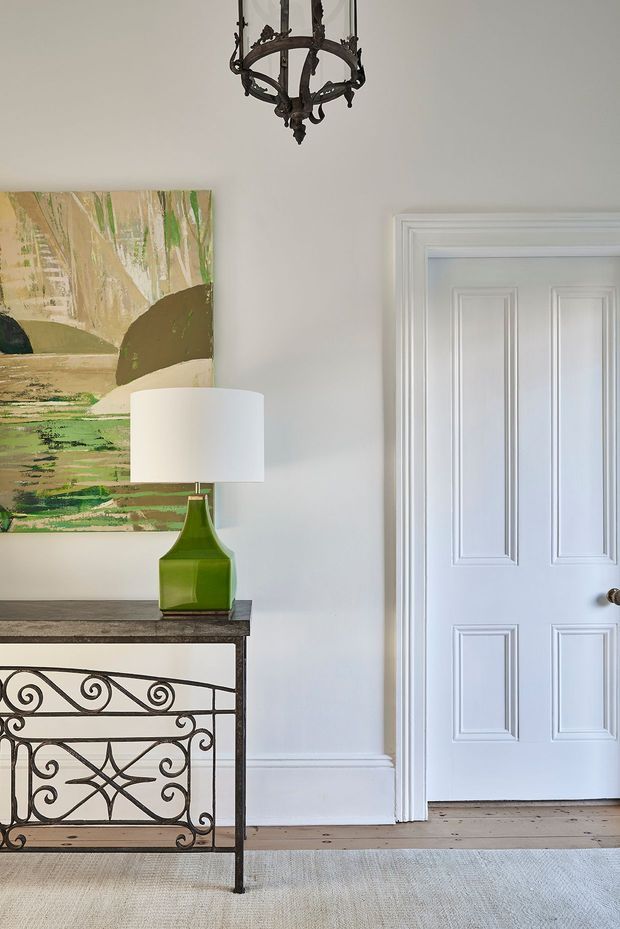 A refined traditional entryway featuring a white paneled interior door with decorative moulded trim, pale neutral walls, a timber floor, and a narrow wrought-iron console table with an ornate scrollwork base. A green glass table lamp, abstract landscape artwork, and an antique-style dark metal chandelier add character.