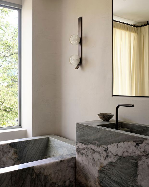 A refined bathroom interior featuring a sculptural freestanding stone bathtub beside a large window, a substantial stone vanity with integrated basin and matte black faucet, a framed mirror, and a vertically mounted two-globe wall light. The space uses dramatic veined natural stone, soft warm plaster walls, and cream curtains, creating a luxurious, moody atmosphere with strong natural light.