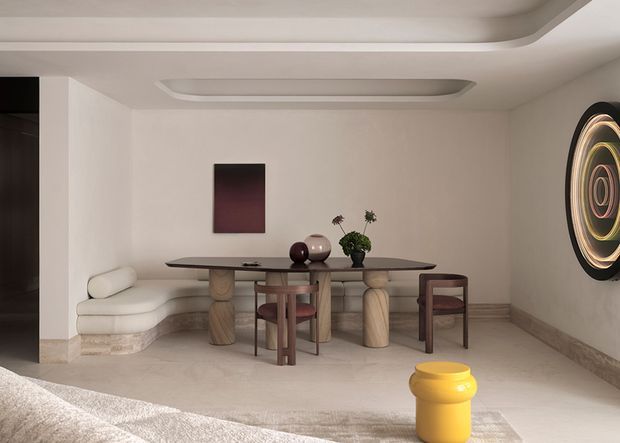 A refined open-plan dining and living area with a sculptural dark dining table, upholstered dining chairs, a curved built-in banquette, and a neutral stone-toned interior. The room features rounded architectural ceiling details, subtle wall panelling or trim, contemporary artwork, and soft sculptural lighting or artwork, creating a calm, luxurious atmosphere.
