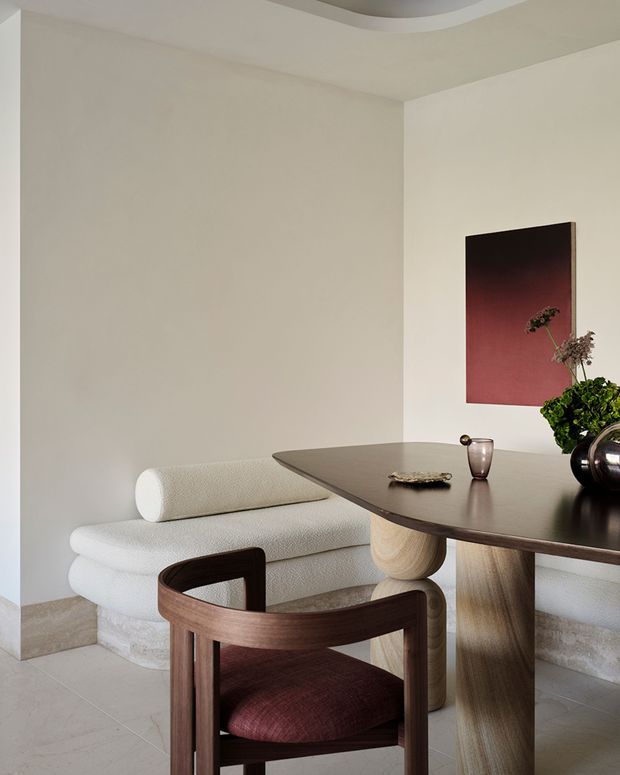 A refined dining corner with a sculptural dark wood dining table and matching wood-framed chair in the foreground. A curved cream boucle banquette sits against warm off-white walls, paired with a rounded bolster cushion. The table has rounded, pedestal-like supports and holds minimalist ceramics and greenery. A dark burgundy abstract artwork adds contrast, while a recessed curved ceiling detail and pale stone-look flooring contribute to the calm, contemporary setting.