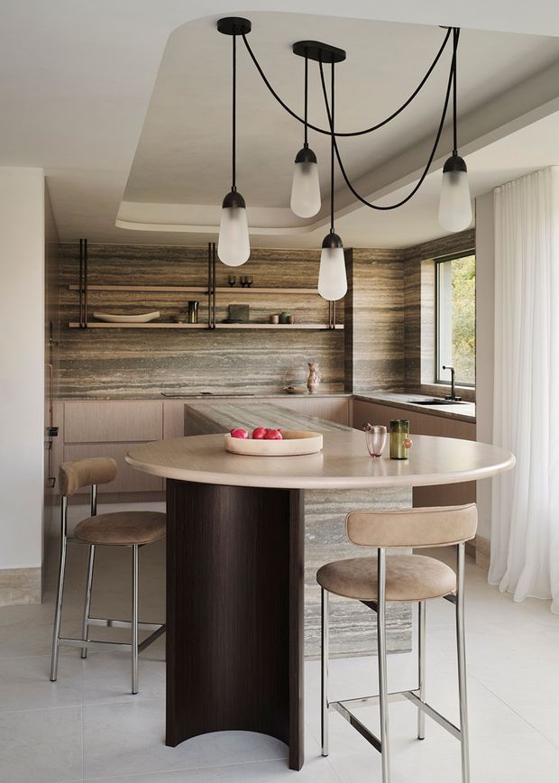 A refined contemporary kitchen and dining area featuring a sculptural rounded island with dark timber cabinetry and a light stone benchtop, surrounded by bar stools. The kitchen has horizontally veined stone wall cladding and splashback, pale timber lower cabinetry, open shelving, integrated appliances, and a large window with sheer curtains. Multiple black-cord pendant lights hang from a recessed ceiling feature, creating a warm, sophisticated atmosphere.