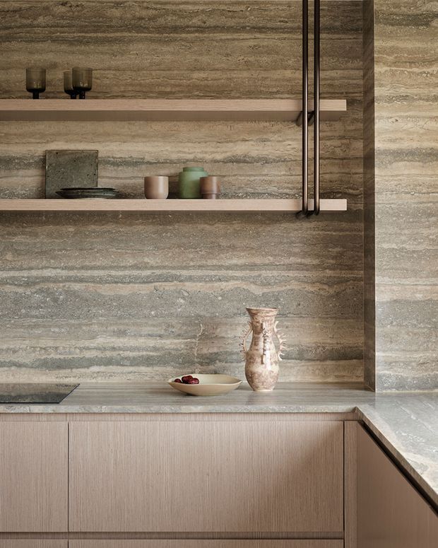 Close-up of a refined contemporary kitchen with a full-height horizontally veined stone backsplash and matching benchtop. Warm taupe vertically textured cabinetry sits below open display shelves holding understated ceramics and glassware. A dark cooktop is partially visible, with integrated vertical metal detailing adding a sculptural accent.