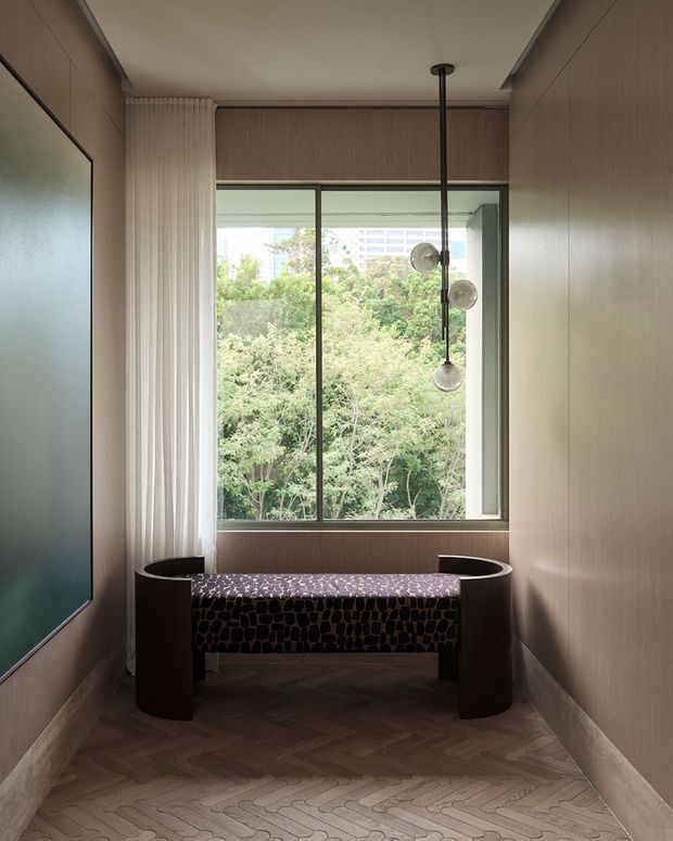 A narrow, refined interior nook with a large framed window overlooking trees, warm wood-panelled walls, a herringbone timber floor, and a sculptural upholstered bench beneath a clustered pendant light. A large framed artwork or screen occupies the left wall, while full-height sheer curtains soften the window. The space has a calm, contemporary and richly textured character.