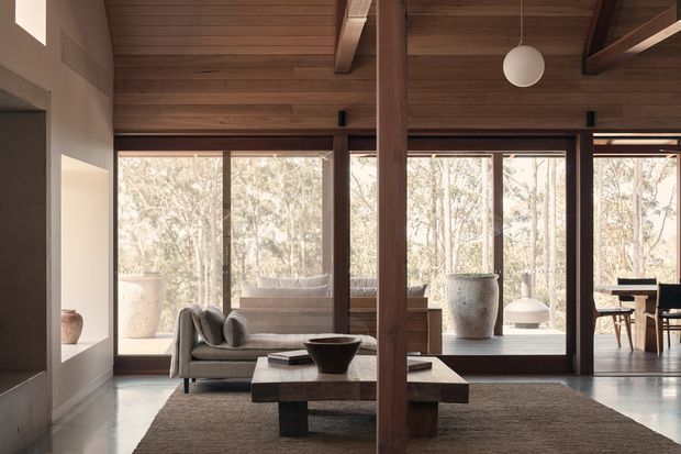 A warm, timber-lined open-plan living space with a low contemporary sofa, substantial timber coffee table, and textured rug. Full-height glazing frames a wooded landscape, while a central timber post and exposed ceiling beams create a strong structural rhythm. A dining area is visible beyond the living room.