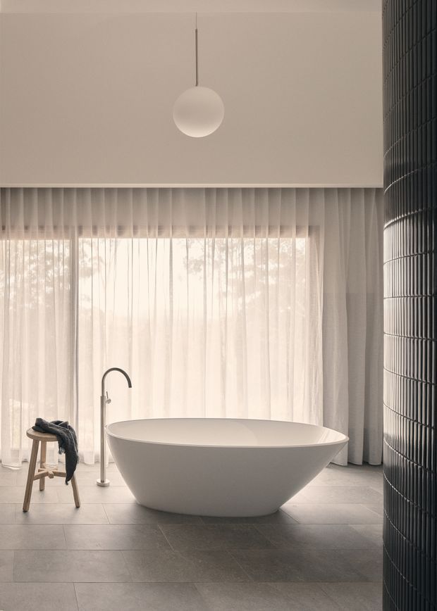 A serene, minimalist bathroom dominated by a freestanding oval bathtub positioned in front of full-height sheer curtains and a large glazed opening. The room has large grey floor tiles, a slim freestanding bath tap, a simple spherical pendant light, and a curved wall or column clad in dark narrow tiles on the right. Soft natural light creates a calm, refined atmosphere.