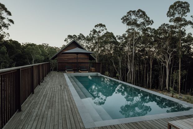 A secluded outdoor pool and timber deck surrounded by dense eucalyptus woodland. The rectangular pool sits flush with the broad deck, which extends toward a small dark timber pavilion with a steep gabled roof and outdoor seating. Dark timber balustrades frame the elevated deck, creating a private bushland retreat with a calm, understated atmosphere.