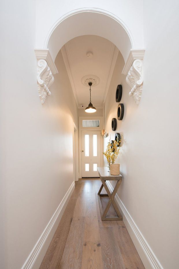 A narrow, elegant residential hallway with warm timber flooring, white walls, decorative architraves, ornate plaster corbels, ceiling mouldings and a central pendant light. A glazed front door is at the far end, with a slim console table, vase of branches and circular wall decor along the right wall.