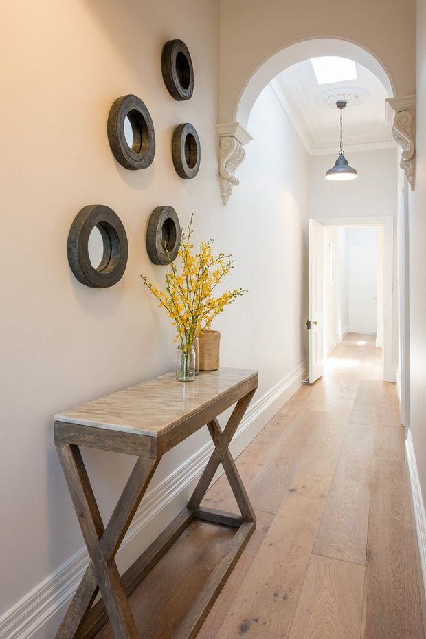 A bright, elegant residential hallway with pale timber flooring, warm off-white walls, detailed white skirting and architraves, and a decorative arched opening framed by ornate plaster corbels. A narrow timber console table with a stone or marble-look top sits against the wall beneath a grouping of dark circular wall mirrors, with a vase of yellow branches. A pendant light hangs beneath a decorative ceiling medallion, and a skylight brightens the corridor.