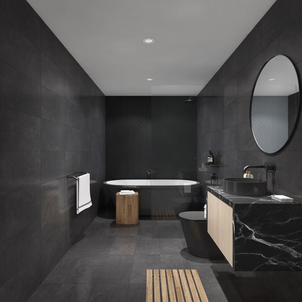 A dark, contemporary bathroom with charcoal wall and floor tiles, a black herringbone feature wall, freestanding bathtub, black vessel basin, wall-mounted black fixtures, round mirror, toilet, and a striking black marble vanity with light timber cabinetry. The space has recessed ceiling lighting and a moody, refined atmosphere.