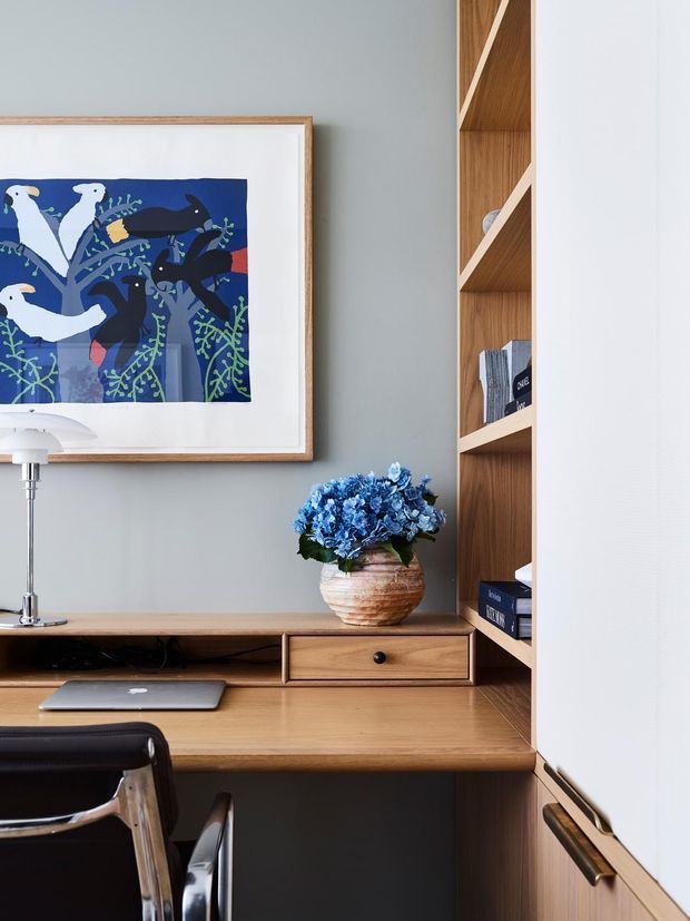 A compact built-in home office nook with a warm timber desk, integrated drawer and open shelving, set against a soft grey wall. A framed blue-toned artwork, desk lamp, laptop, books, and a blue floral arrangement add colour and character. The joinery continues into white cabinetry with brass-toned hardware.