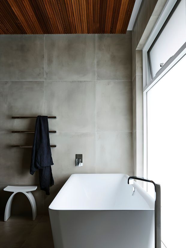 A modern bathroom featuring a freestanding white bathtub positioned beside a large corner window. The walls are clad in large grey concrete-look panels, complemented by a warm timber slatted ceiling. A dark towel hangs on minimalist wall-mounted rails, with a small white stool nearby. The restrained palette and clean geometric forms create a calm, refined atmosphere.