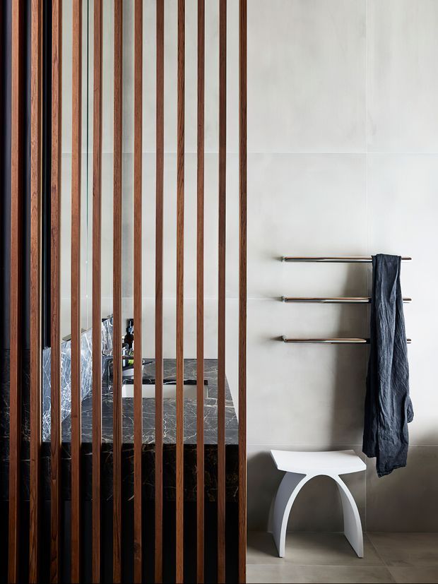 A refined contemporary bathroom featuring a freestanding dark stone bathtub partially screened by slim vertical timber battens. Large pale wall tiles, dark tiled flooring, stainless-steel heated towel rails, and a sculptural white stool create a clean, high-contrast interior.