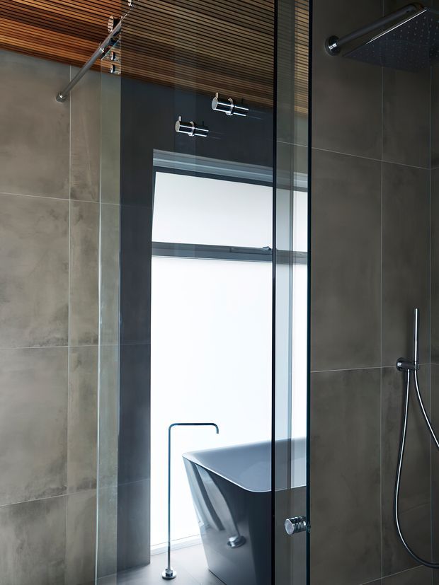Modern bathroom interior featuring a frameless glass shower enclosure, large grey stone-look wall tiles, a freestanding dark bathtub, chrome fixtures, and a warm slatted timber ceiling. A high window provides strong natural light.