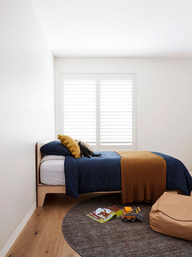 A compact, bright child's bedroom with white walls and ceiling, a timber bed frame, navy bedding, and a mustard knitted throw. White plantation shutters cover the central window. A round dark rug, tan beanbag, children's books, and a toy construction vehicle add casual playfulness to the room.