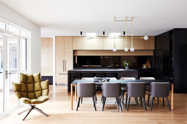 Bright open-plan contemporary kitchen and dining area with pale timber cabinetry, a dark island and back wall, a long dining table with upholstered chairs, and floor-to-ceiling glazing along one side. A sculptural mustard lounge chair and linear pendant lighting add contrast to the restrained interior.