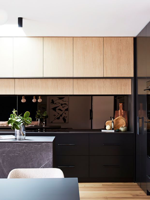 A contemporary kitchen viewed from the dining area, featuring full-height pale timber overhead cabinetry, matte black lower cabinetry, dark reflective splashback, and a dark stone island with a light benchtop. Warm timber flooring, pendant lights, indoor plants, and restrained styling create a refined, moody interior.