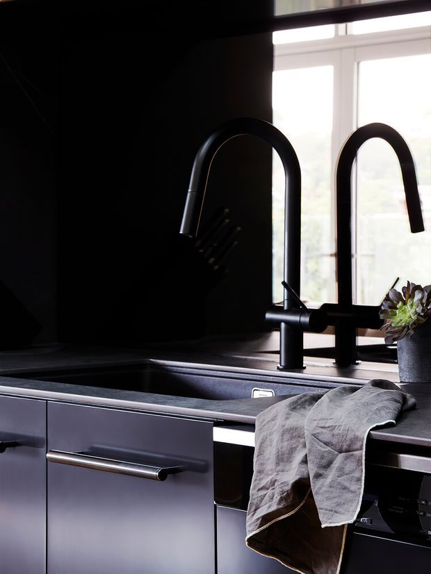 Close-up of a dark, contemporary kitchen sink area with matte black gooseneck faucets, a deep rectangular sink, dark benchtops, and sleek grey cabinetry with long metal handles. A large white-framed window provides bright natural light, while a potted plant and linen tea towel add subtle contrast.