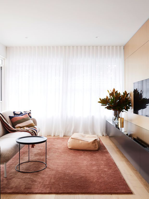 Bright contemporary living room with floor-to-ceiling sheer white curtains, pale timber wall panelling and flooring, a wall-mounted television, low dark media cabinetry, a blush-pink rug, round black coffee table, sofa with a patterned throw, and a soft ottoman. The room has abundant natural light and a warm, refined neutral palette.
