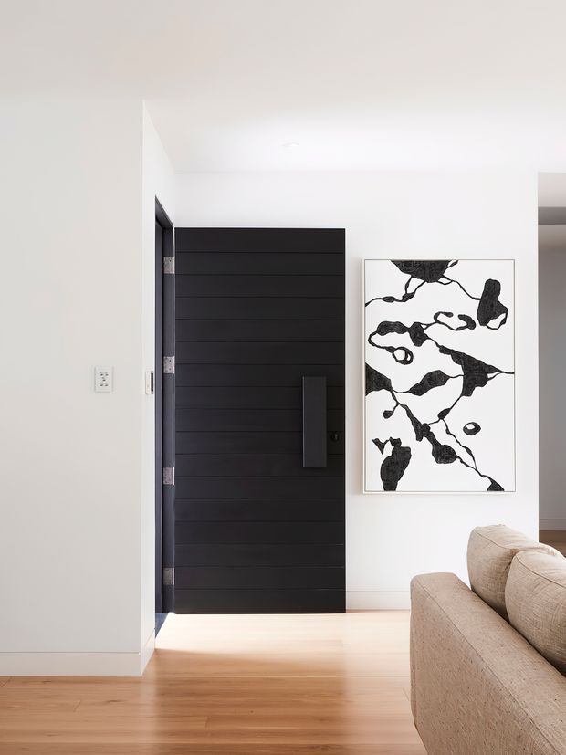Bright minimalist residential entry area with a tall black horizontally paneled front door, white walls, light timber flooring, and a beige upholstered sofa partially visible in the foreground. A monochrome abstract artwork hangs beside the door.