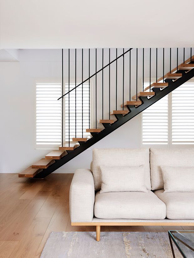Bright contemporary interior featuring a minimalist open-riser staircase with a black steel stringer and slim vertical balustrade wires, paired with warm timber treads. The staircase spans a light-filled living room with white plantation shutters, pale timber flooring, and a large cream upholstered sofa.