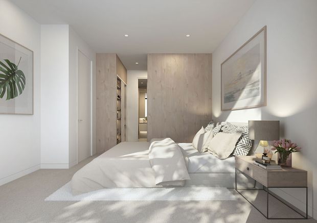 Bright, calm contemporary bedroom with a large upholstered bed, neutral bedding, pale timber wall and wardrobe joinery, light carpet, framed artwork, and a slim bedside table. A hallway and built-in storage are visible beyond the bed, while recessed ceiling lights and soft daylight create an airy, refined atmosphere.