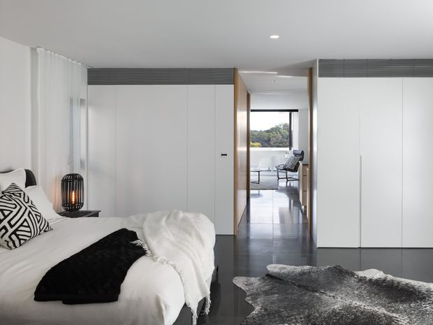 A bright, minimalist bedroom with a white upholstered bed, patterned monochrome cushions, a black throw, bedside table and pendant lamp. Floor-to-ceiling white built-in wardrobes line the room, accented by dark horizontal upper panels. A polished dark floor leads through an open passage to a naturally lit sitting area beyond, while sheer curtains cover the left-side glazing.