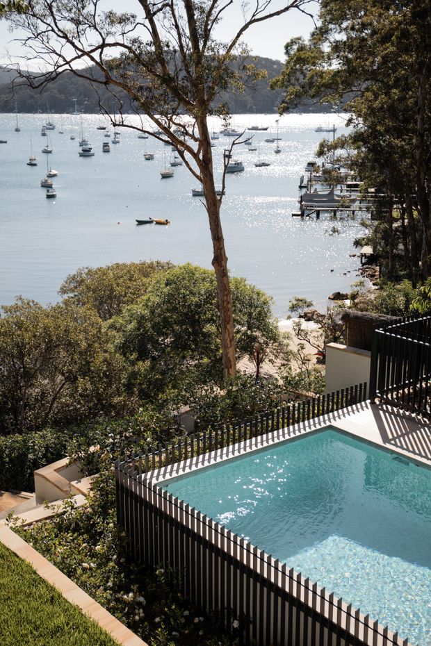 A bright elevated outdoor view overlooking a calm, boat-filled bay, framed by mature trees and landscaped vegetation. In the foreground is a modern rectangular swimming pool with pale blue water, bordered by dark vertical metal fencing, light paving, and stepped outdoor terraces. A small jetty and shoreline are visible beyond the property.