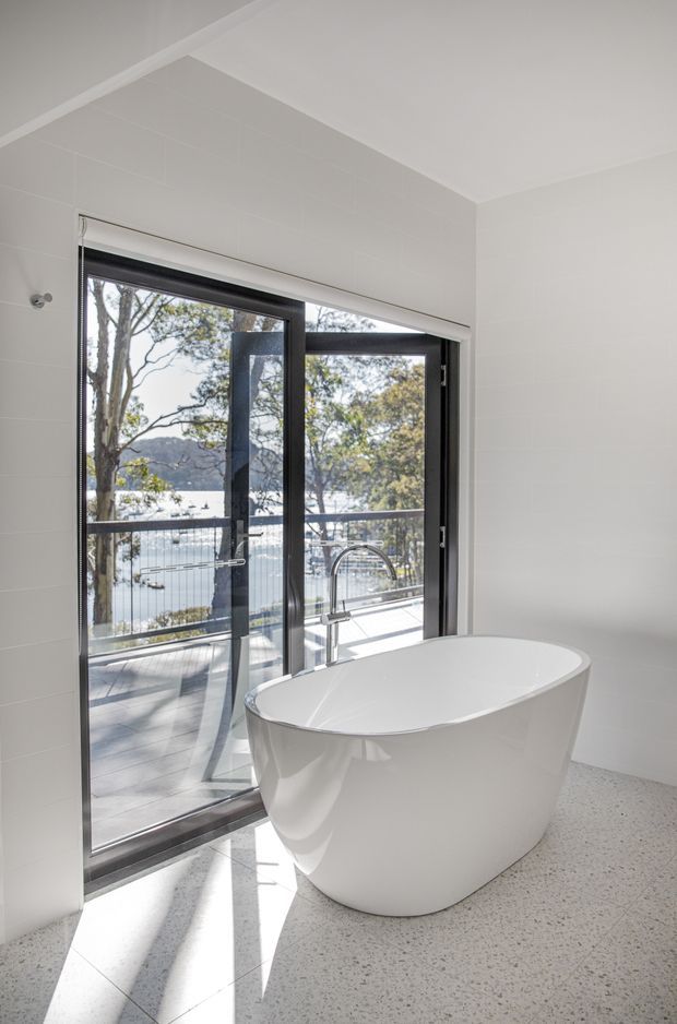 Bright contemporary bathroom with a freestanding white oval bathtub positioned beside full-height dark-framed glazing and a balcony or terrace overlooking a body of water. White tiled walls, a pale terrazzo-style floor, and a chrome floor-mounted bath filler create a clean, airy setting.