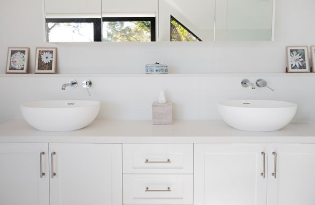 Bright, symmetrical contemporary bathroom vanity with two white vessel basins set on a pale stone or solid-surface benchtop. The vanity has white built-in cabinetry with brushed metal handles, wall-mounted chrome taps, and a large mirror spanning the wall above. Small framed botanical artworks and a tissue box sit on the counter, while windows and greenery are reflected in the mirror.