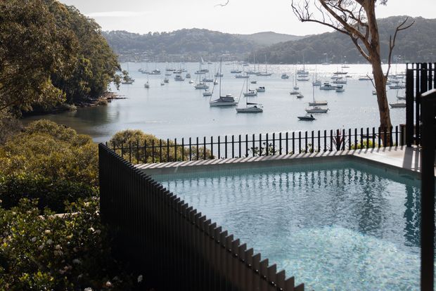 A tranquil waterfront view from a raised outdoor terrace, featuring a rectangular swimming pool enclosed by black vertical metal fencing. Beyond the pool, a sheltered bay is filled with moored sailboats, framed by dense coastal vegetation, mature trees, and forested hills. The scene has a refined contemporary outdoor character with strong black-and-water-blue contrast.