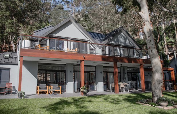 A contemporary multi-level home set among mature bushland, with two prominent pitched gabled roof forms, dark metal roofing, light rendered walls, extensive glazing, and a broad timber-framed upper balcony. The lower level opens to a covered outdoor living area overlooking a lawn, with a large eucalyptus tree in the foreground.