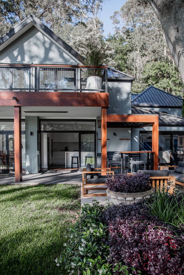 A contemporary multi-level house set among mature trees, viewed from a landscaped garden. The facade combines pale rendered walls, dark standing-seam metal pitched roofs, extensive black-framed glazing, and warm reddish-brown timber framing and balcony elements. A covered outdoor terrace with dining furniture opens toward a lawn and layered garden planting.