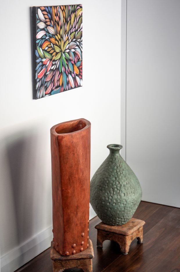 A styled interior corner with white walls, dark timber flooring, and a closed white door. Two large decorative vessels stand on small rustic wooden plinths: a tall reddish-brown cylindrical vessel and a rounded green textured vase. Above them hangs a colourful abstract botanical artwork with radiating leaf-like forms.