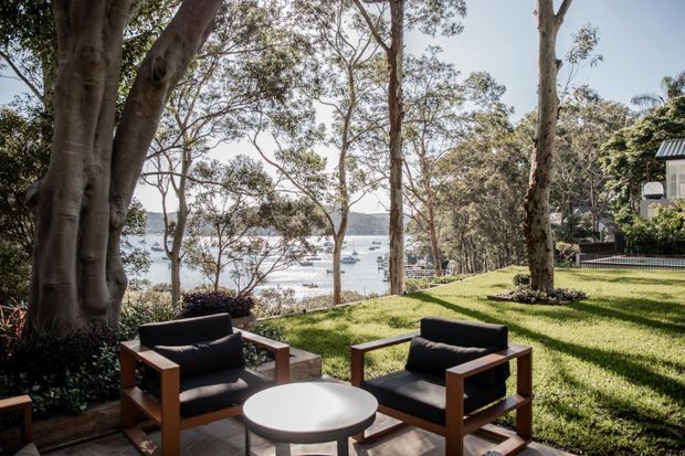 A landscaped waterfront outdoor sitting area beneath mature trees, furnished with two timber-framed armchairs and a round coffee table. The lawn slopes gently toward a calm bay with boats visible through the trees, while part of a modern residential building appears at the right edge.