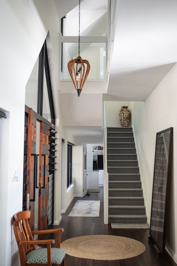 A bright, narrow residential entry hall viewed from inside, featuring a dark timber floor, white walls, a carpeted staircase with a small landing, and a mezzanine or upper-level void with glass-panel balustrading. A sculptural wood-and-metal pendant hangs above the circulation space. On the left is a substantial dark timber and glass entry door or screen with adjacent glazing, while a timber armchair, framed artwork, console/display elements, and layered round and rectangular rugs add warmth. The hallway extends toward a bright living area at the rear.