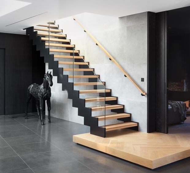 Contemporary interior view dominated by a sculptural open-riser staircase with black zigzag steel stringers, timber treads, and a clear glass balustrade. A pale timber handrail runs along a light grey textured wall, contrasting with dark timber wall panelling and large grey floor tiles. The staircase rises from a raised timber platform beside a dark doorway leading to a partially visible bedroom. A black horse sculpture stands beneath the stairs.