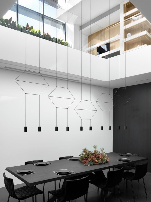 A dramatic double-height contemporary dining area with a long dark dining table, black woven chairs, sculptural geometric pendant lights, white walls, and dark timber wall cladding. Floor-to-ceiling glazing and an upper-level void with planted greenery and warm timber shelving create a bright, refined architectural interior.