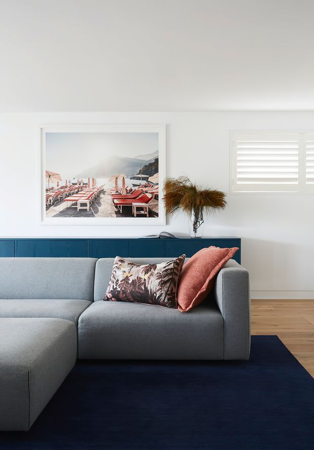 A bright, refined living room featuring a large light-grey sectional sofa, navy rug, deep blue built-in cabinetry, pale timber flooring, white walls, plantation shutters, and coastal artwork. Coral and patterned cushions add colour, while a small sculptural dried-grass arrangement sits on the cabinetry.